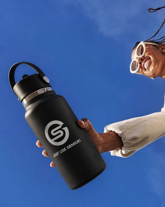 A smiling person wearing sunglasses and a beige sweater holds up a black Hydro Flask-style water bottle printed with a logo and the text 'JUST USE GENSUKI' against a bright blue sky.
