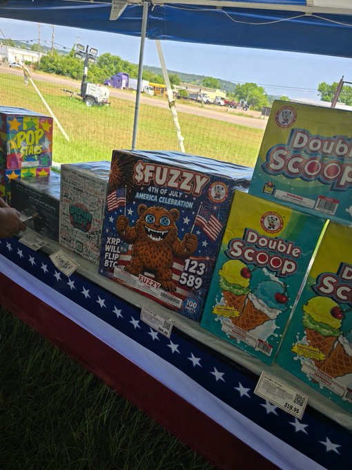 A fireworks stand display features "SEUZZY" branded fireworks celebrating the "4th of July American Celebration" with XRP Will Flip BTC messaging, alongside "Double Scoop" and other fireworks boxes, tying into the post's $FUZZY/XRP Independence Day theme.