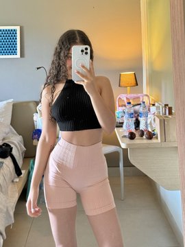 Mirror selfie of a woman wearing a black halter top and pink ribbed biker shorts, standing in a room with a vanity table, lamp, and bed in the background.