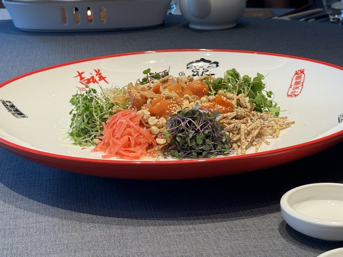 A vibrant Asian salad featuring microgreens, pickled ginger, shredded chicken, and diced salmon is artfully plated on a red-rimmed dish decorated with Chinese characters, presented at what appears to be a Binance compliance event.