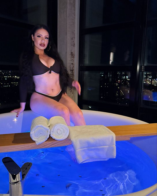 A woman poses confidently in a black bikini beside a glowing bathtub with neatly rolled towels, set against a backdrop of city lights at night. The luxurious setting complements the post's motivational message about stepping out of one's comfort zone to pursue dreams.