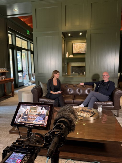 A behind-the-scenes podcast setup shows two people seated on a brown leather couch in a green-walled room, with a professional camera in the foreground and its monitor displaying the live recording feed.
