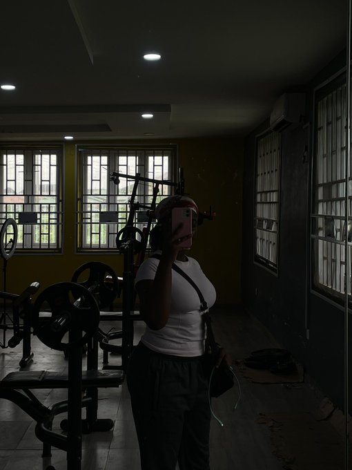 A woman takes a mirror selfie in a dimly lit gym, surrounded by weight racks and weight plates, capturing her first to-do list item of hitting the gym. The atmospheric lighting through the large windows highlights her white tank top and casual athletic fit, embodying the start of her productive day.