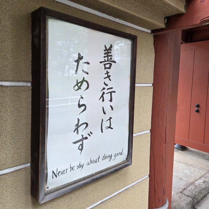 ikebana art style flower arrangement on a beige wall, accompanied by Japanese calligraphy and the phrase "Never be shy about doing good."