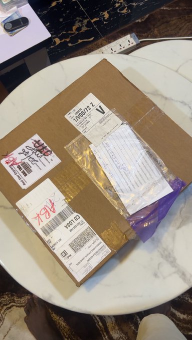 A cardboard shipping package sits on a marble table, displaying visible shipping labels with tracking numbers "VN089722" and "0227146" along with a customs document, likely containing Cronos App branded merchandise.