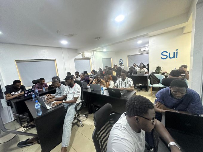 A group of developers works intently on laptops during the Walrus Sessions Hackathon, with "Sui" branding visible on the wall behind them. The image captures the collaborative, in-person coding event referenced in the post, showcasing attendees like @Glo_rious9 engaged in building with Walrus Memory technology.