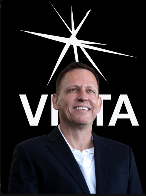A smiling man in a dark suit and white shirt poses in front of a black background featuring a white star logo and the letters 'VE' and 'TA' partially visible behind him.