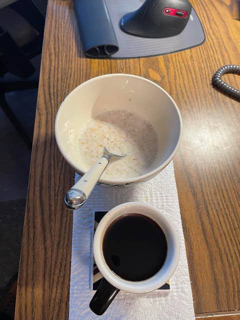 A nearly empty cereal bowl with a spoon sits next to a full cup of black coffee on a wooden desk.