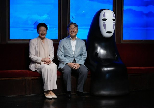 Two people sitting on a red bench next to a statue of the No-Face character from the movie Spirited Away, in front of windows showing a blue sky.