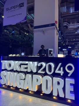 A large 3D sign reading 'TOKEN2049 SINGAPORE' is displayed at an event venue, with a person working at a counter in the background and a 'SUPER AI' banner hanging above.