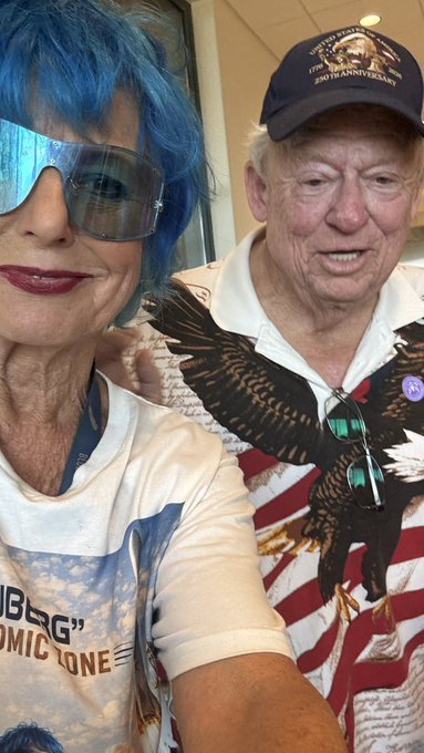 An older woman with bright blue hair and large sunglasses poses for a selfie alongside an older man wearing a 'United States of America 250th Anniversary' commemorative cap and an eagle-patterned shirt.