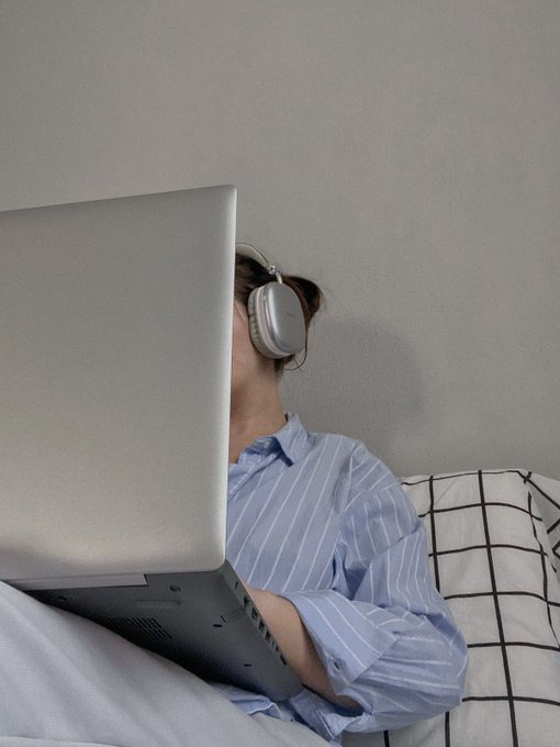 The image shows a person wearing headphones while lying down with their head resting on a pillow. They are using a laptop computer that is placed on their lap, suggesting they might be working or browsing the internet. The background of the photo appears to be a wall, which could indicate that this scene takes place in someone's bedroom or living space.