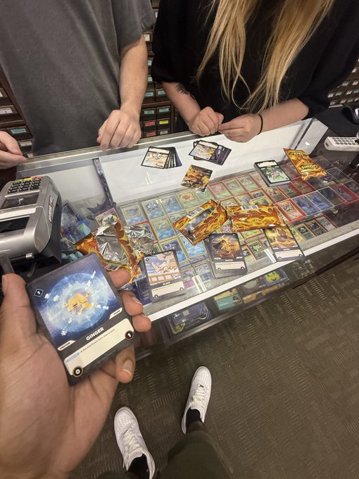 First-person view of someone holding a Pokemon trading card at a store counter, with a display case of various cards and three other people visible across the glass counter.