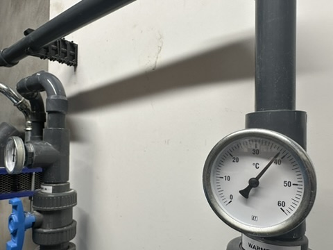 A photograph of a circular analog thermometer mounted on industrial PVC piping, showing a reading around 40°C.
