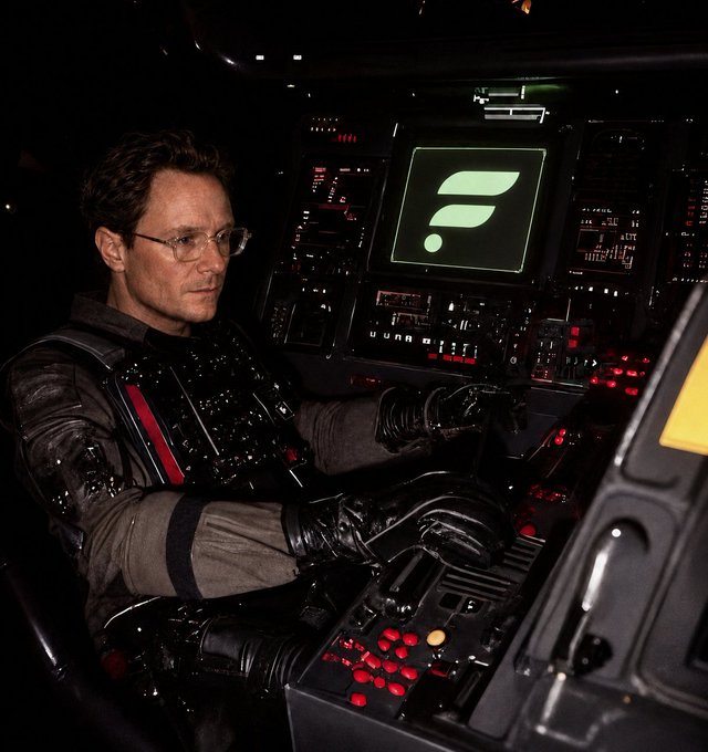 A man wearing glasses and dark tactical gear sits at the glowing control console of a futuristic cockpit or command center, intently operating levers and buttons while a green radar-like symbol pulses on the central monitor. The image's high-stakes, focused atmosphere of piloting or commanding a complex system visually reinforces the post's directive "LOCK IN."—evoking the intense concentration needed to commit fully to a mission or goal.