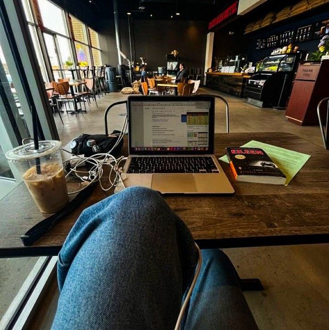 A relaxed first-person view of someone working at a coffee shop with a MacBook displaying spreadsheets, an iced coffee, and a book titled "THE ALCHEMIST" on the table. The image visually reinforces the post's scenario of casually reviewing an AI-generated market summary over coffee while tracking crypto trends.