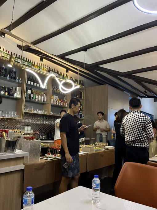 A photograph of the interior of a modern bar or lounge with several people standing around, featuring a neon 'Voo' sign, shelves stocked with liquor bottles, and a water bottle on a white table in the foreground.