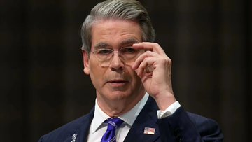 Treasury Secretary Scott Bessent, wearing a suit and glasses, adjusts his glasses while speaking at a formal event.
