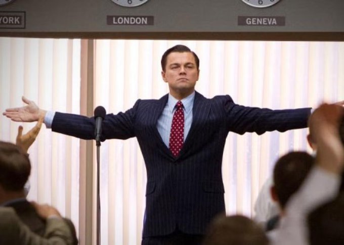Leonardo DiCaprio portrays Jordan Belfort in "The Wolf of Wall Street," arms outstretched like a stockbroker rallying his boiler room team, with city labels "LONDON" and "GENEVA" (and partial "YORK") displayed above — the iconic scene the post references to contrast old-school penny stock manipulation with $PRM's modern ticker-listing model that eliminates the need for such operations.