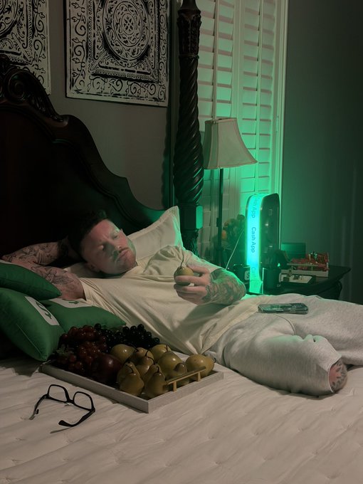 A man reclines in bed looking at his phone, with glasses, a tray of fruit, and a glowing green “Cash App” sign beside him. The scene matches the post’s playful “BAAANG @CashApp We in here! #CashAppPartner” message by highlighting the Cash App partnership.