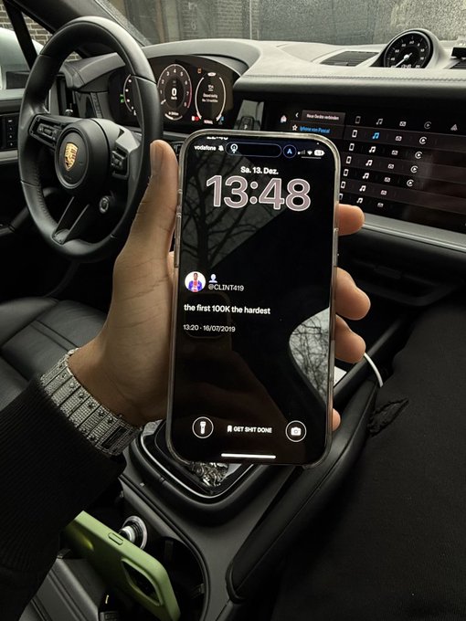 A hand holds up an iPhone inside a Porsche, displaying the lockscreen time 13:48 on Saturday, 13 December, along with a motivational message "the first 100K the hardest" from @CLINT419—visually echoing the post's optimistic sentiment about reaching the $100K milestone in the crypto market.