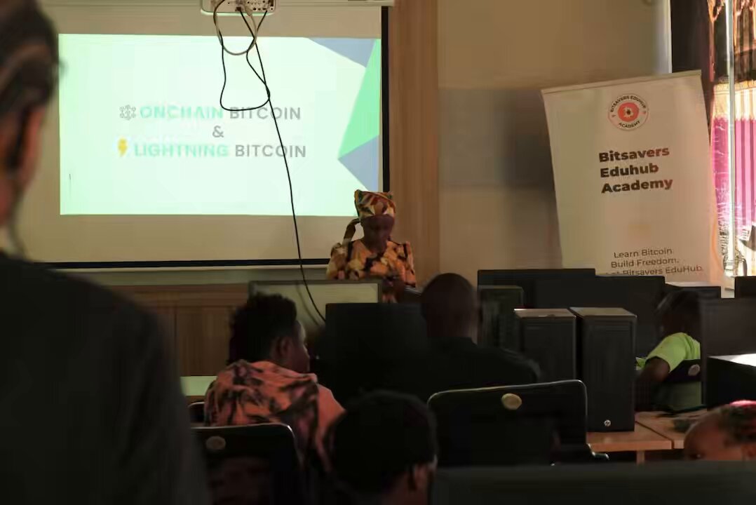 The image shows a group of people sitting in chairs, with one person standing behind a screen displaying information about Bitcoin. The room appears to be a classroom setting where attendees are learning about Bitcoin and its uses.