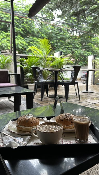 A café tray with two bagel sandwiches, a cappuccino, and a latte glass, set against a lush green garden view through the window.