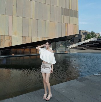 A photo of a woman in a white off-shoulder top and white skirt standing on a paved area near a modern building with a water feature and stone steps.