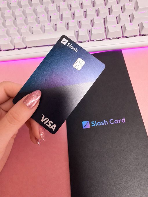 A hand holds up a sleek dark blue Slash Visa credit card above its accompanying packaging labeled "Slash Card," with a white backlit keyboard and pink surface visible in the background.