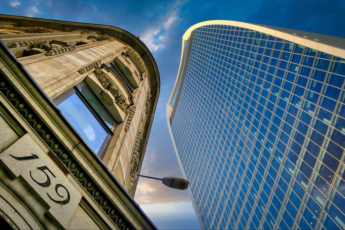 An upward-angled architectural photograph contrasting an ornate classical stone building with the year '1899' inscribed on it against a modern glass-and-steel skyscraper, both meeting against a blue sky with scattered clouds.