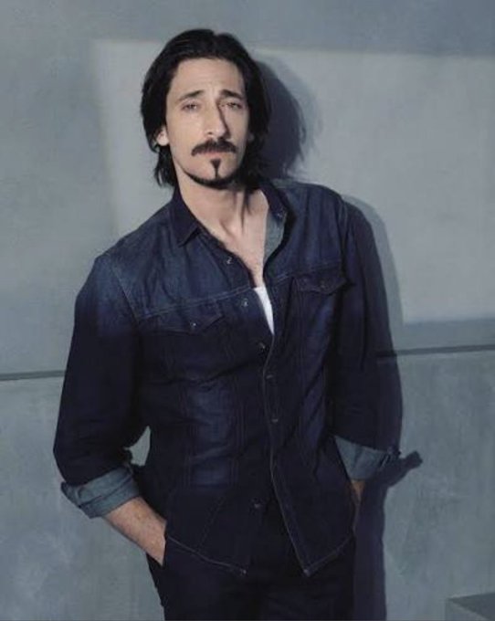 A man with shoulder-length dark hair, a mustache, and a goatee stands in a dark denim jacket against a gray background.