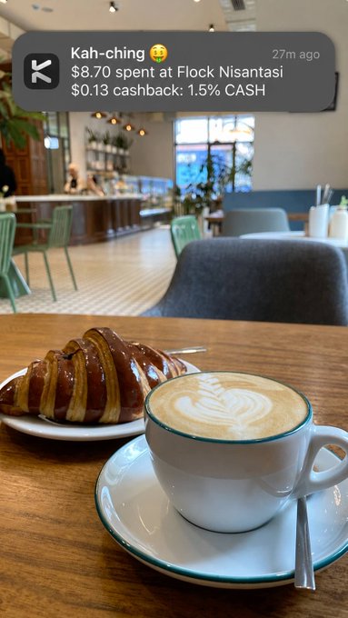 A café scene showing a cappuccino with latte art and a chocolate-glazed croissant on a wooden table, with a notification overlay reading "Kah-ching 😄 $8.70 spent at Flock Nisantasi $0.13 cashback: 1.5% CASH" from 27m ago.