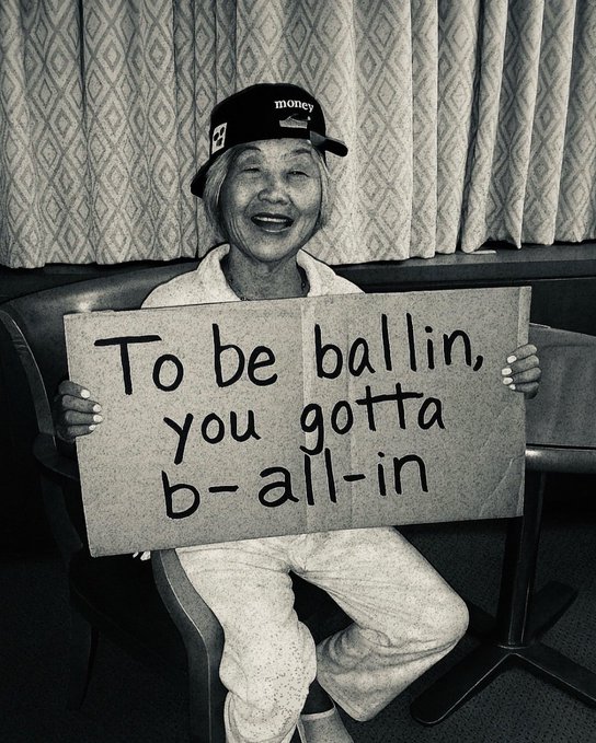 The image features an older woman sitting down, wearing a baseball cap with the number "3" on it. She is holding up a sign that reads "To be ballin' you gotta b-all-in". The background of the photo shows a chair and a dining table, suggesting that she might be in a casual setting like a restaurant or a gathering place where people can sit down and enjoy their time together.