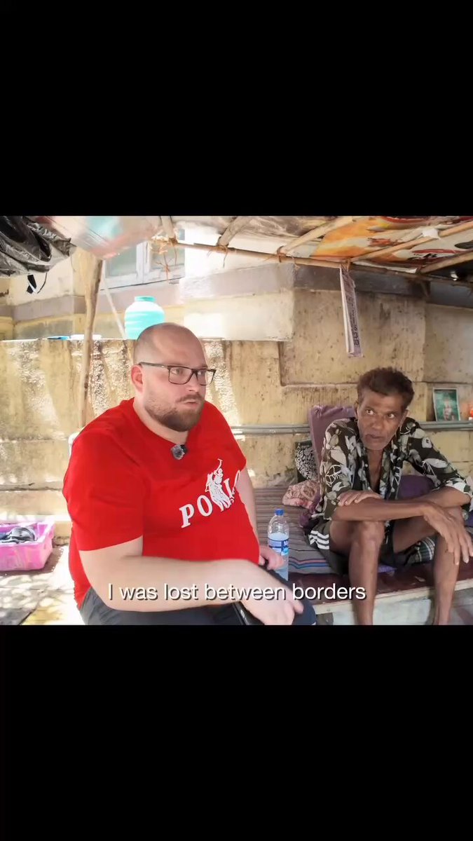 There are two men sitting next to each other, one wearing a red shirt and another wearing a green shirt with black writing that appears to be from a video game. In the background, there is a truck.