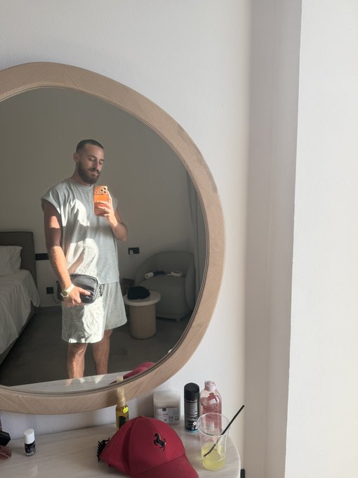 A man with a beard takes a mirror selfie in a hotel room, holding an orange phone with "BURGER" visible on the case, while wearing a grey t-shirt and holding a black pouch. The "Vibe check" caption fits the casual, relaxed aesthetic of the minimalist room, with a red cap and drinks on the vanity below.