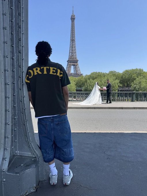 A person stands on a bridge facing the Eiffel Tower, wearing a black shirt with yellow 'CORTEL' lettering on the back, baggy jeans, and white sneakers, while a couple poses for wedding photos nearby.