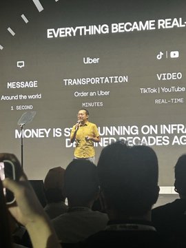A man in a yellow shirt gives a presentation on stage in front of a screen displaying 'EVERYTHING BECAME REAL' along with diagrams about Uber, TikTok, YouTube, transportation, and messaging services, with audience members visible in the foreground.