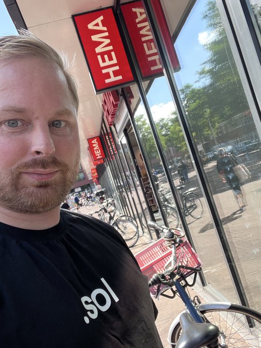 A selfie of a bearded man wearing a black t-shirt with '.sol' printed on it, taken outside a HEMA store with bicycles visible in the window.