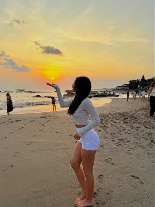 A young woman in white shorts and a long-sleeve top stands on a sandy beach at sunset, appearing to catch the sun in her hand, with other people visible along the shoreline.