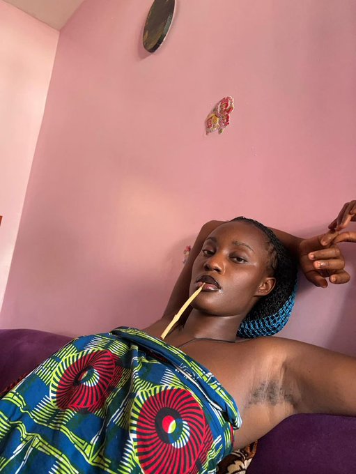 A woman reclines confidently against a pink wall, wearing vibrant Ankara fabric and a blue headwrap, holding a slender stick near her lips in a poised manner. The caption "My bride price is 2BTC" pairs with her self-assured pose, humorously presenting herself as a valuable "commodity" with a cryptocurrency price tag, playing on modern dating and transactional themes.