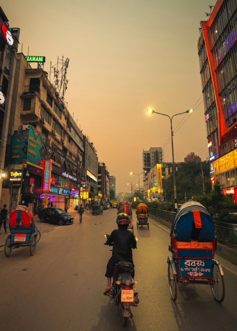 The image shows a busy city street with people riding motorcycles and rickshaws, as well as cars driving down the road. The sky appears hazy, suggesting that the photo was taken during sunset or early morning hours. A person wearing a helmet is riding a motorcycle in front of a building on the left side of the frame. In addition to this individual, there are several other people visible throughout the scene, some walking and others driving their vehicles down the street. The image also includes text that reads "good night my x family, for surviving every chapter that tried to break me @NomismaNetwork See you in the morning @wallchain @quipnetwork".