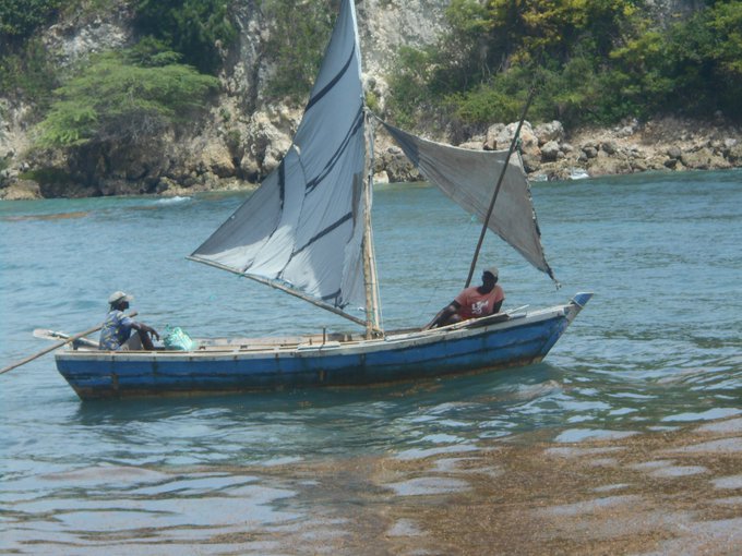 A photograph of two men in a small blue wooden sailboat with tattered white sails on bright blue waters, with a rocky, tree-lined shore in the background.
