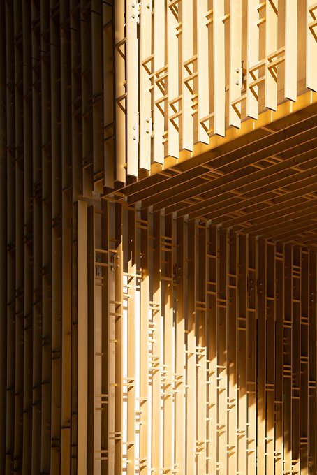An architectural detail showing wooden slats arranged in a geometric pattern with triangular cutouts, lit with warm sunlight creating dramatic shadows.