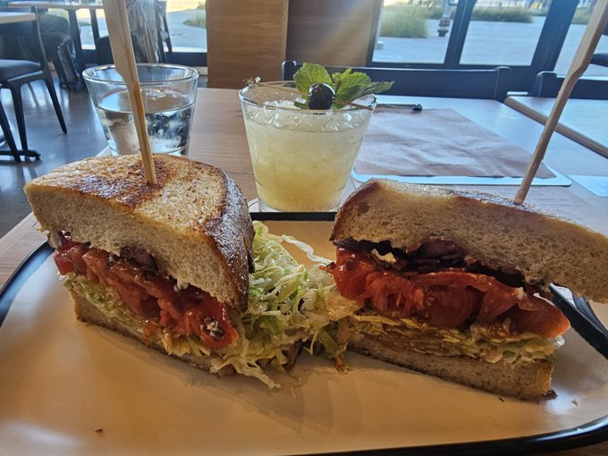 A club sandwich with sun-dried tomatoes, cheese, and lettuce is served on a white plate alongside a refreshing cocktail garnished with mint and a blackberry, fitting the celebratory vibe of "Cheers degens." The drink and hearty sandwich make for a satisfying post-worthy meal.