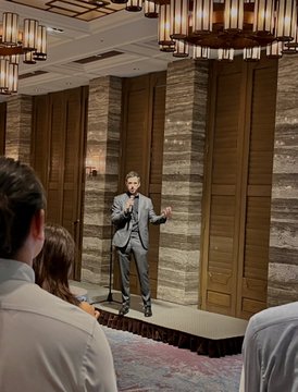 A man in a gray suit speaks on stage in an elegant venue with ornate chandeliers and wood-paneled walls, appearing to give a presentation or talk. The image shows the speaker mentioned in the post sharing a moment on stage with audience members visible in the foreground, consistent with the live speaking engagement described.