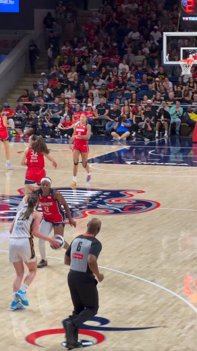 The image shows a basketball game with two teams playing against each other, one wearing red jerseys and the other wearing white. The players are actively engaged in the game, running up and down the court as they compete for points. In the background, there is an audience watching the match intently from their seats. A referee can be seen standing near them, ensuring that the game proceeds fairly and according to the rules.