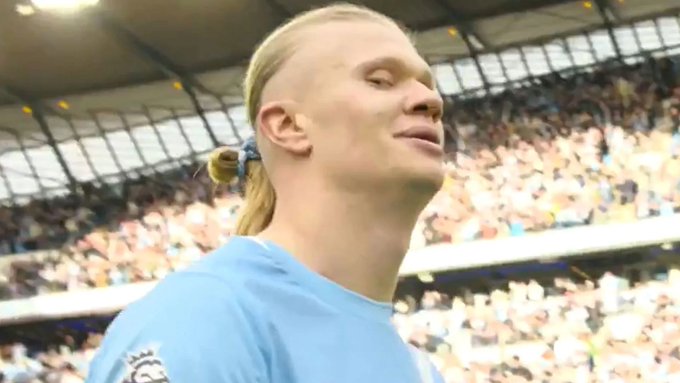 Football player Erling Haaland wearing a light blue Manchester City jersey, smiling and looking upward with his signature long blonde hair tied back.