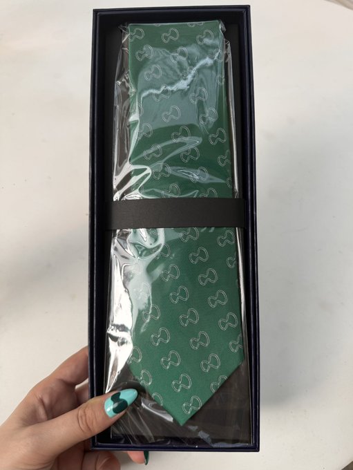 A hand holds an open black box containing a green necktie with a horseshoe pattern, still sealed in clear plastic packaging.