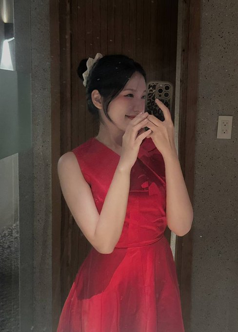 A young woman in an elegant red dress with a floral hair accessory takes a mirror selfie with her smartphone in a softly lit room with wooden walls.