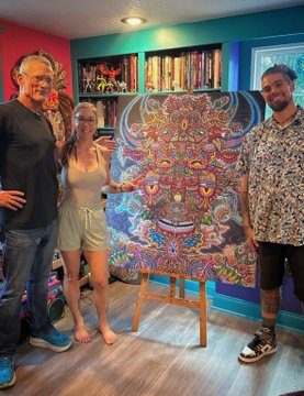 Three people pose around a colorful, intricate psychedelic painting displayed on an easel in a vibrant room with bookshelves.